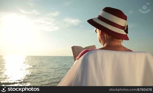 Senior woman relaxing with a good book by sea on the beach at sunset, rear view