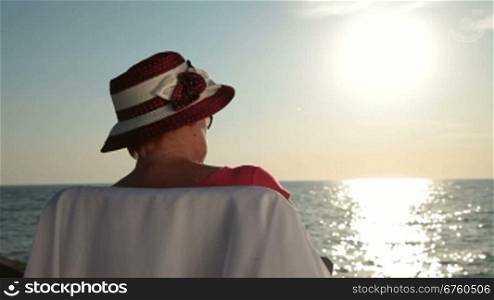 Senior woman relaxing with a good book by sea on the beach at sunset, rear view