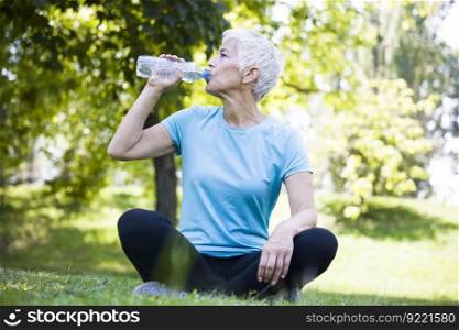 Senior woman sitting and drinking water after workout in park