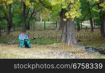 Senior woman sitting outdoors in chair under autumn tree, reading book Front View