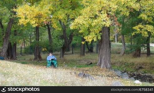 Senior woman sitting outdoors in chair under autumn tree, reading book. Front View