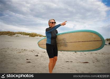 Senior woman standing on beach, holding surfboard, pointing to sea