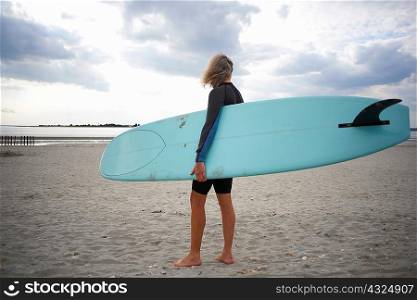 Senior woman standing on beach, holding surfboard, rear view