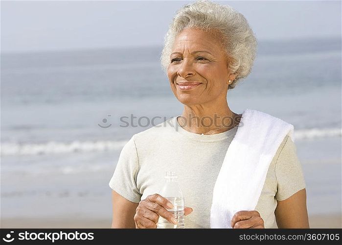 Senior woman stands with drinking water and towel over her shoulder on beach