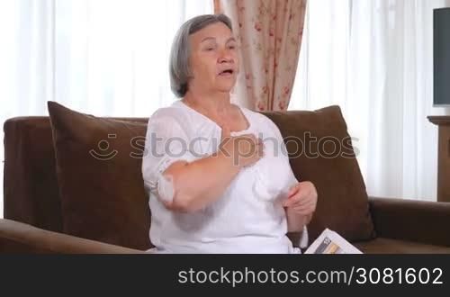 Senior woman suffering from heartache or heart attack while she reads the newspaper at home. Old age, health problem and people concept. Slow motion