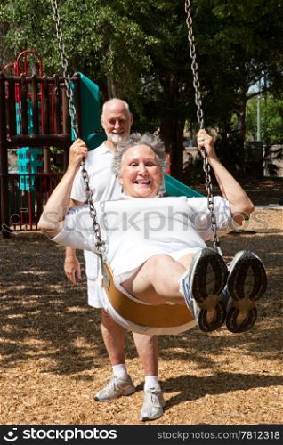 Senior woman swinging on the playground in the park. Her husband is pushing her.