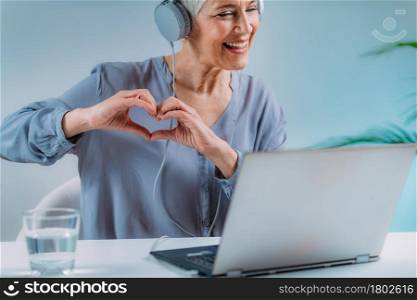 Senior woman using a laptop, having a video call with loved ones and sending love, heart hand sign.. Senior Woman. Video Call with Loved Ones, Sending Love.