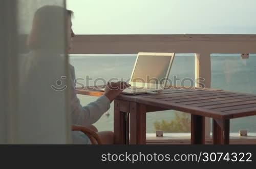 Senior woman using laptop sitting at the table on outdoor balcony with view to the sea. She surfing online using mobile internet device connected through usb