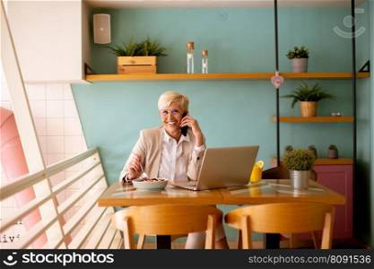 Senior woman using mobile phone while working on the laptop and having healthy breakfast in the cafe