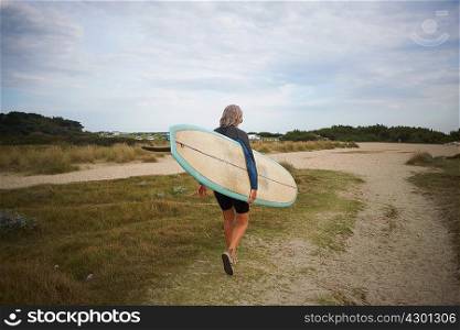 Senior woman walking along beach, carrying surfboard, rear view