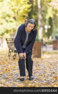 Senior woman walking in autumn park and having knee pain. Arthritis pain concept.