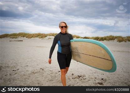 Senior woman walking on beach, carrying surfboard