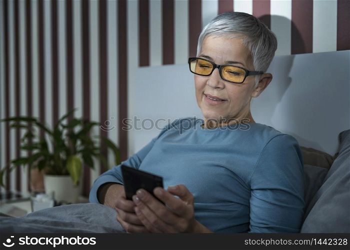 Senior woman with blue-light blocking glasses lying in bed before sleeping, using smartphone.. Senior Woman with Blue Light Blocking Glasses Using Smartphone in the Evening