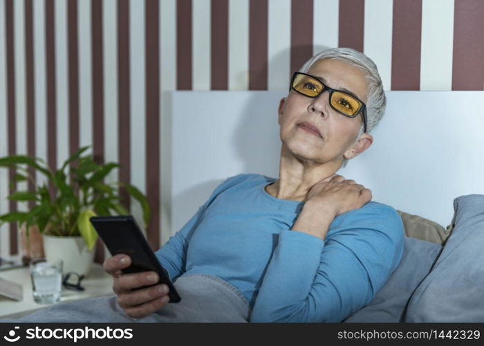 Senior woman with blue-light blocking glasses lying in bed before sleeping, using smartphone.. Senior Woman with Blue Light Blocking Glasses Using Smartphone in the Evening