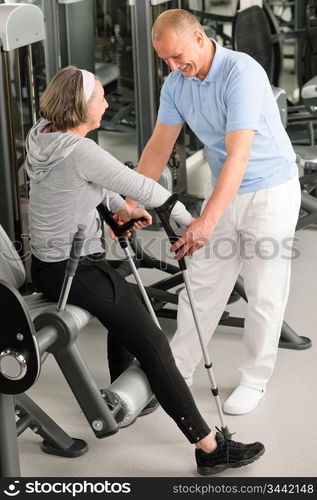 Senior woman with crutches getting help of physiotherapist at gym