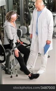 Senior woman with crutches getting help of physiotherapist at gym