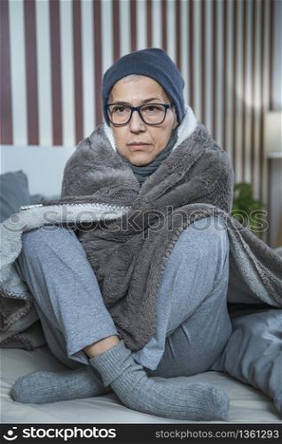 Senior woman with symptoms of seasonal flu, covered with blanket, sitting on the bed at home, feeling ill.. Senior Woman with Symptoms of Seasonal Flu