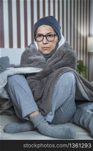 Senior woman with symptoms of seasonal flu, covered with blanket, sitting on the bed at home, feeling ill.. Senior Woman with Symptoms of Seasonal Flu