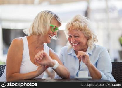 Senior women laughing in street cafe. Two happy mature women in outdoor cafe. One is using smart watch while another looking at it
