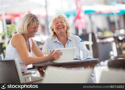Senior women laughing while using tablet PC in cafe. Two female friends using touch pad in street cafe and laughing. Senior women spending enjoyable summer day