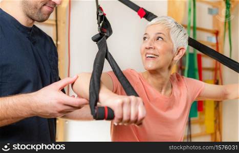 Senior women training arms with trx fitness straps in the gym with coach&rsquo;s help. Senior Woman Exercising on TRX