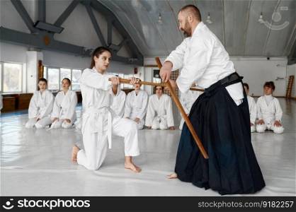 Sensei with young female student fighting with wooden sword demonstrating aikido skills to group of teenage woman fighters. Sensei with young female student fighting with wooden sword
