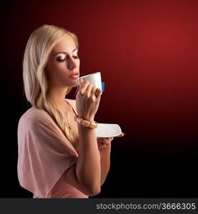 sensual blond girl with hair style drinking a cup of tea in elegant pink dress and bracelet over dark fashion background