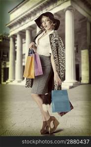 sensual blonde girl with kitsch leopard coat and big black hat posing with colorful shopping bags. in vintage color