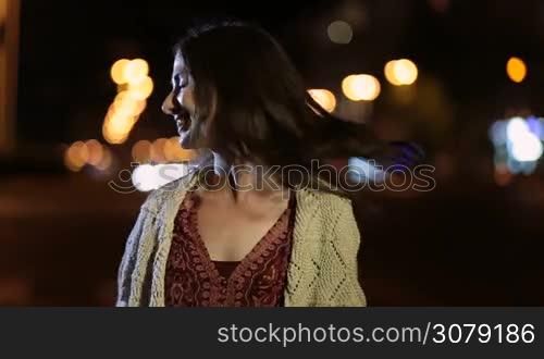 Sensual brunette female with long hair posing on night city street and smiling playfully. Sexy young woman in stylish clothes flirting with camera at night over passing by traffic and streelights background. Slow motion. Steadicam stabilized shot.