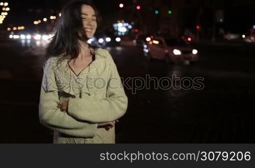 Sensual brunette female with long hair posing on night city street and smiling playfully. Sexy young woman in stylish clothes flirting with camera at night over passing by traffic and streelights background. Slow motion. Steadicam stabilized shot.