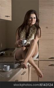 sensual brunette woman sitting in kitchen with sexy winter short dress and a cup of coffee in the hand. She is looking in camera