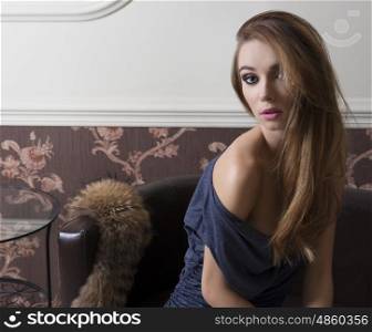 sensual elegant woman with blue dress is sitting on leather sofa near fur. sensual fashion woman with make-up, she is looking in camera with lovely expression