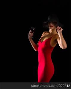 sensual girl in a bright red dress and black wide-brimmed hat stands with a glass of wine on a dark background. girl hat and wine
