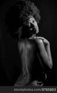 Sensual nude African American female with curly hair looking away over shoulder while standing against black background. Black and white photograph.. Black and white photograph of naked black woman under neon light