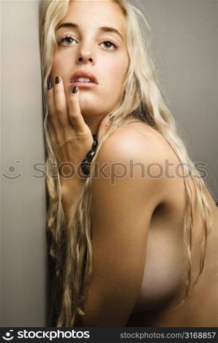 Sensual nude Caucasian woman leaning on wall with hand to face.