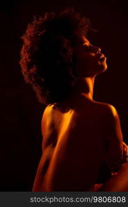 Sensual topless African American female model embracing body and closing eyes under orange neon illumination against black background.. Calm black woman embracing naked torso