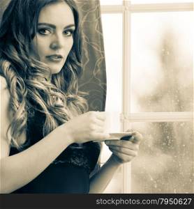 Sensual woman drinking hot coffee beverage at home. Sensual seductive woman in lingerie drinking cup of coffee by curtain and french door window at home. Young girl with hot energizing beverage stay awake. Caffeine energy. Black and white.