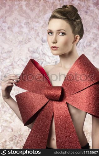 sensual woman with elegant blonde hair-style posing in christmas shoot with big red glitter bow covering her naked breast, looking in camera