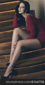 sensual woman with long black hair, sexy red dress and cute legs is sitting on elegant stairs and looking in camera