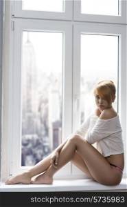 sensual woman with red hair in sexy pose near window with naked legs , panties and sexy t-shirt looking in camera