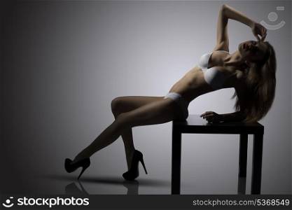 sensual young brunette girl posing with her perfect body on small table wearing sexy white lingerie and heels. Backlight glamour portrait