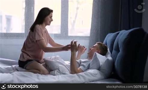 Sensual young couple looking at each other passionately on bed in morning. Attractive couple in love spending leisure together in the bed after awake. Pretty brunette female riding on boyfriend and fooling around. Side view.