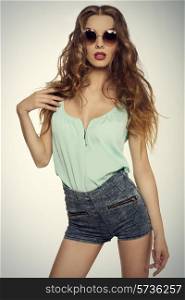 sensual young female with long curly hair and red lipstick wearing hipster sunglasses sexy blue shirt and denim shorts. Looking in camera, slim body