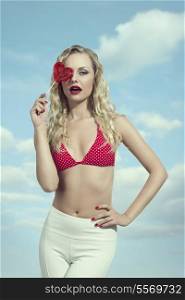 sensual young woman with curly blonde hair-style posing with heart shaped valentines lollipop and wearing bikini and white pants