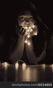 Sepia toned portrait of a young attractive woman with long hair sitting on the floor, in a dark room, holding in her hands a string of lights.