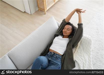 Serene african american girl napping, relaxing on comfortable sofa at home, enjoying break, lazy weekend. Relaxed young female stretching arms, resting, lying on couch in cozy living room.. Serene african american girl napping, relaxing lying on comfortable sofa at home, stretching arms
