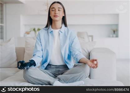 Serene disabled woman is sitting in lotus pose on couch. Lady practicing morning yoga with her eyes closed. Attractive european girl has myoelectric cyber prosthesis. Modern artificial limb.. Serene disabled woman sitting in lotus pose on couch practicing morning yoga with her eyes closed.