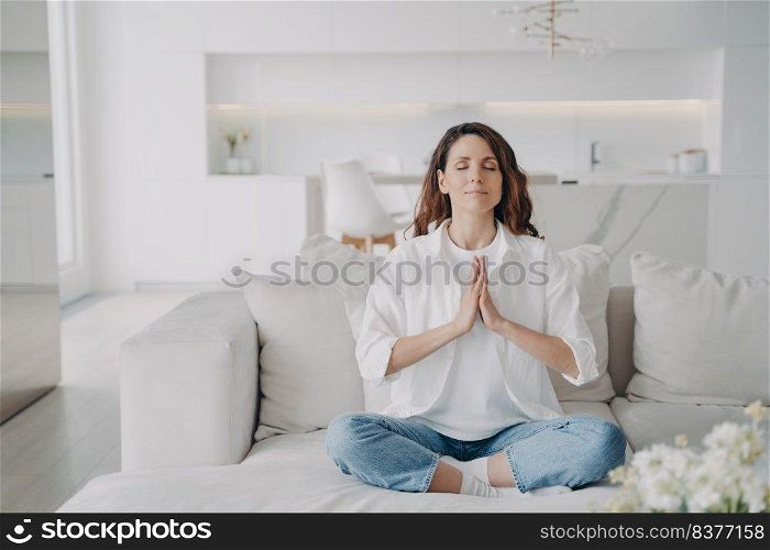 Serene latin woman at home. Posture exercise, lotus asana. Young european woman is practicing yoga and meditation on couch with her legs crossed. Minimal white modern scandinavian interior.. Serene young latin woman practicing yoga and posture exercise, lotus asana at home.