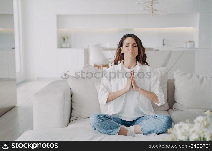 Serene latin woman at home. Posture exercise, lotus asana. Young european woman is practicing yoga and meditation on couch with her legs crossed. Minimal white modern scandinavian interior.. Serene young latin woman practicing yoga and posture exercise, lotus asana at home.