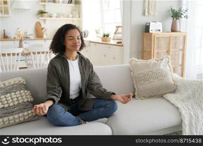 Serene mixed race girl meditates, does yoga, sitting on couch in lotus pose at home. Calm young lady enjoying meditation, does breathing exercises on sofa to relieve stress. Healthy lifestyle concept.. Serene mixed race girl meditates does yoga sitting on couch in lotus pose at home. Healthy lifestyle